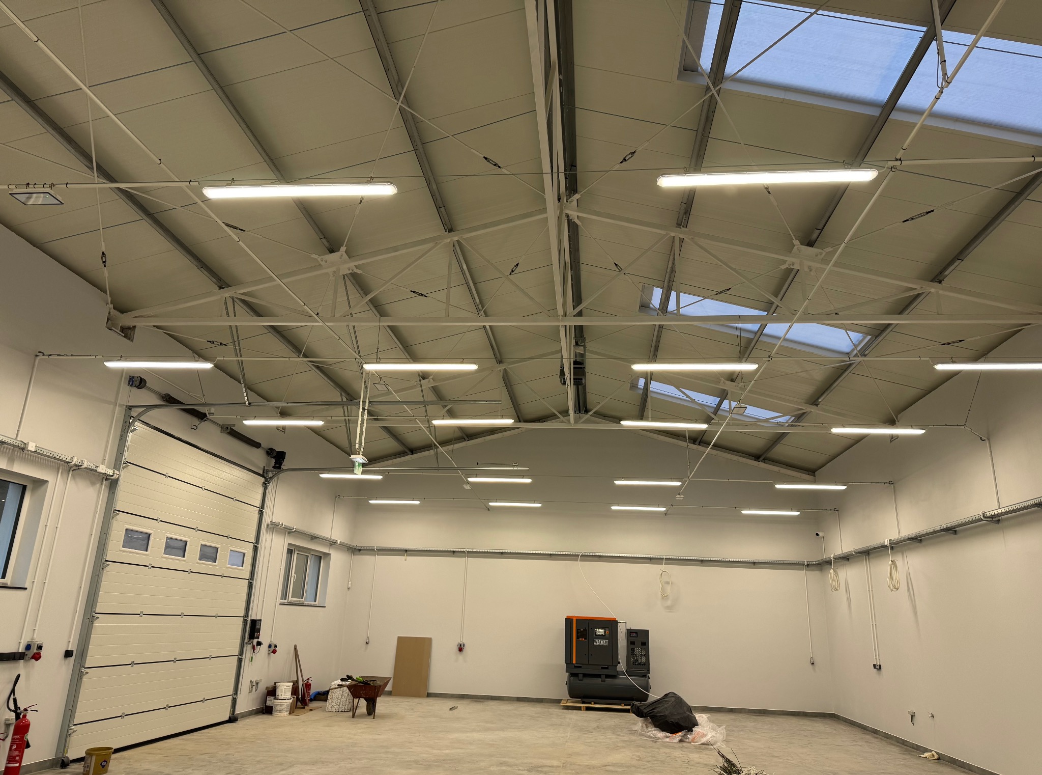 Wnętrze hali z zamontowanym oświetleniem LED. Widoczna brama segmentowa, białe ściany i sufit z widoczną konstrukcją. Dobre oświetlenie przestrzeni.