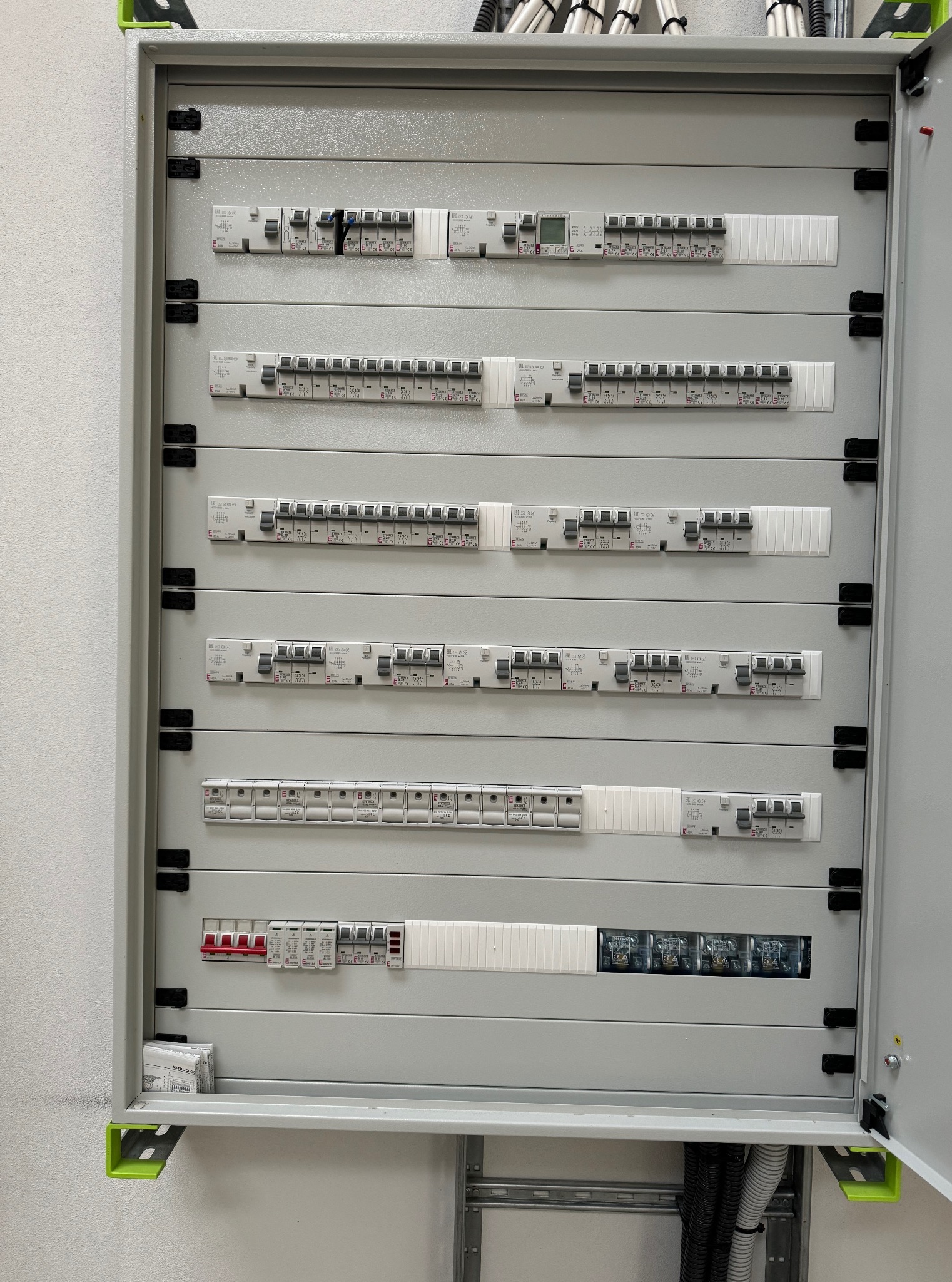 Rozdzielnica elektryczna z bezpiecznikami, szyny DIN i okablowanie wewnątrz szafy. Widoczne elementy instalacji elektrycznej, szare tło, oświetlenie sufitowe.