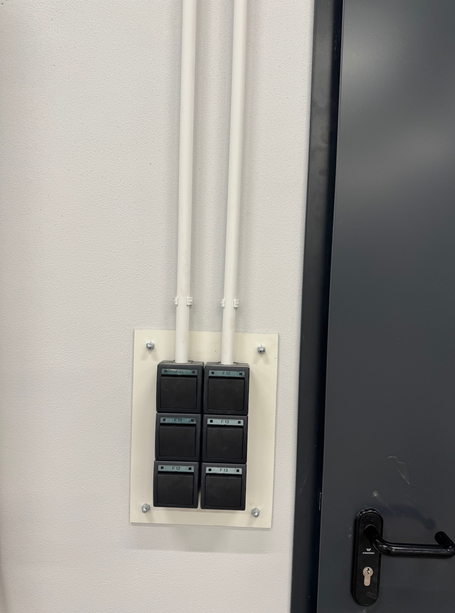 Panel sterowania z sześcioma czarnymi przyciskami (F10-F13) na białej płycie, przymocowany do ściany. Nad nim dwie białe rurki. Obok fragment szarych drzwi.
