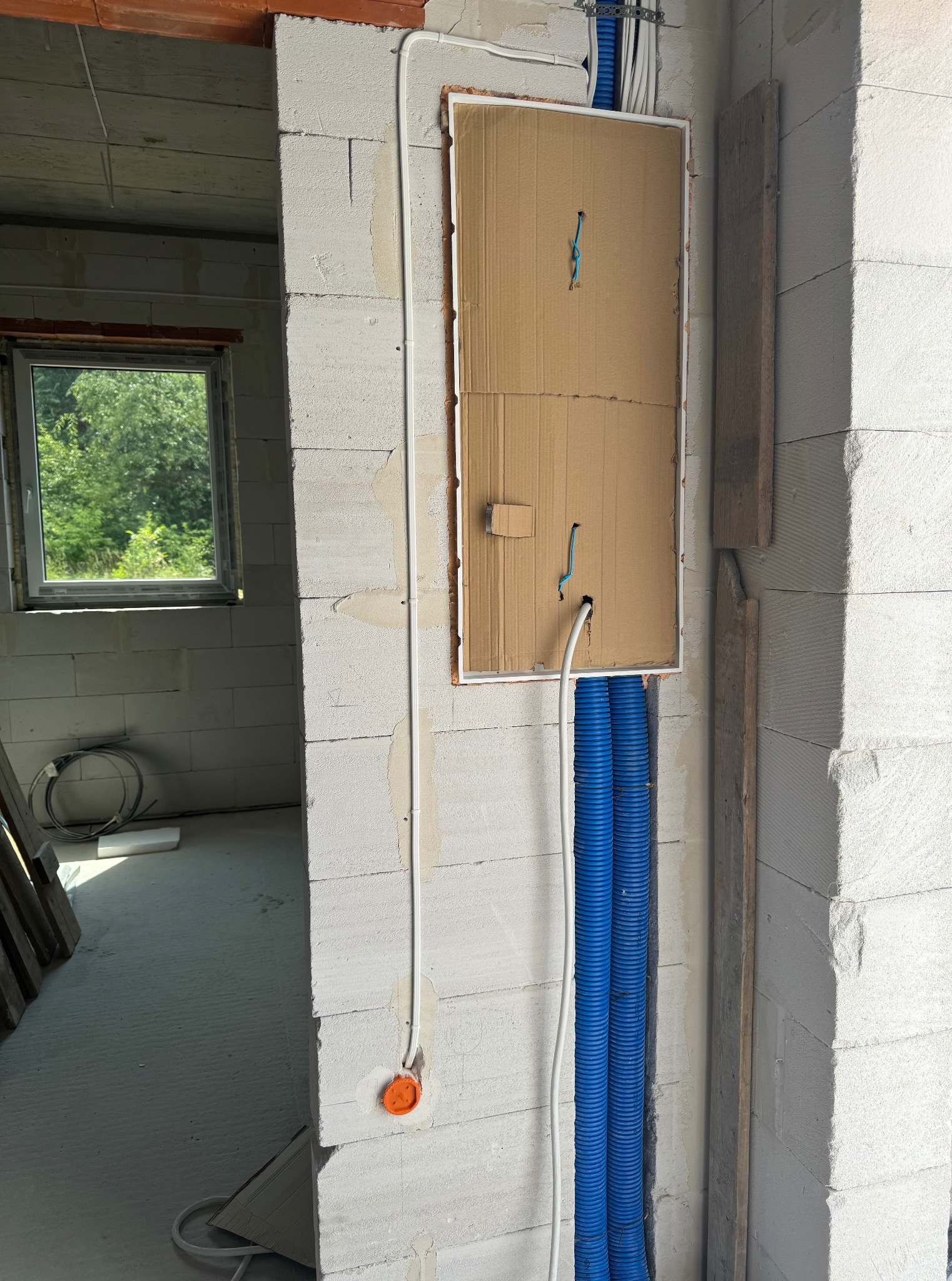 Instalacja elektryczna w surowym wnętrzu: białe ściany z betonu komórkowego, niebieskie rury karbowane, przewody i puszka elektryczna. Widok na okno i zieleń na zewnątrz.