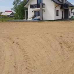 Wyrównany teren pod budowę domu jednorodzinnego. Widoczne ślady po maszynach niwelacyjnych. W tle dom z balkonem i tarasem. Słoneczny dzień.