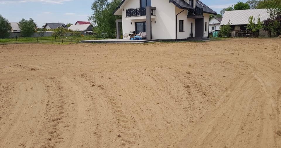 Wyrównany teren pod budowę domu jednorodzinnego. Widoczne ślady po maszynach niwelacyjnych. W tle dom z balkonem i tarasem. Słoneczny dzień.