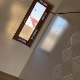 Sebastian Ślusarczyk - Biała łazienka z oknem dachowym w drewnianej ramie, wykończona błyszczącymi płytkami z trójkątnym wzorem na ścianie. Widok na zewnątrz przez okno.