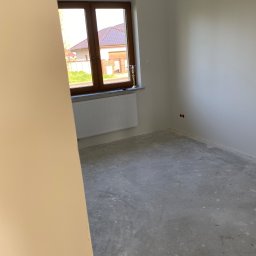 Sebastian Ślusarczyk - Surowe wnętrze pokoju z oknem, widoczne ściany pomalowane na jasny kolor, betonowa podłoga i grzejnik pod oknem. Gotowe do dalszych prac wykończeniowych.