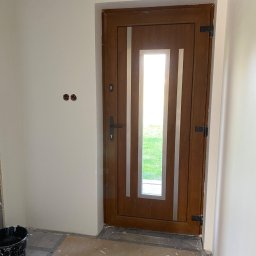 Sebastian Ślusarczyk - Brązowe drzwi wejściowe z szybą, osadzone w świeżo pomalowanej, jasnej ścianie. Widoczne ślady prac wykończeniowych na podłodze i puszki elektryczne bez osprzętu.