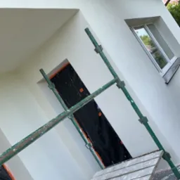 Biała elewacja domu po renowacji w Szprotawie, widoczna z poziomu rusztowania. Okno i otwór zabezpieczone folią malarską.