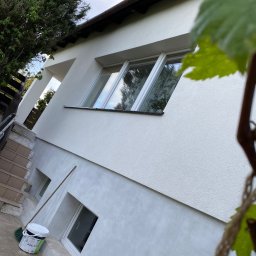 Sebastian Ślusarczyk - Ocieplony, biały budynek z widocznymi oknami, z boku widoczny pęd winorośli. Przy ścianie stoi wiadro z farbą i miotła. Świeżo pomalowana elewacja.