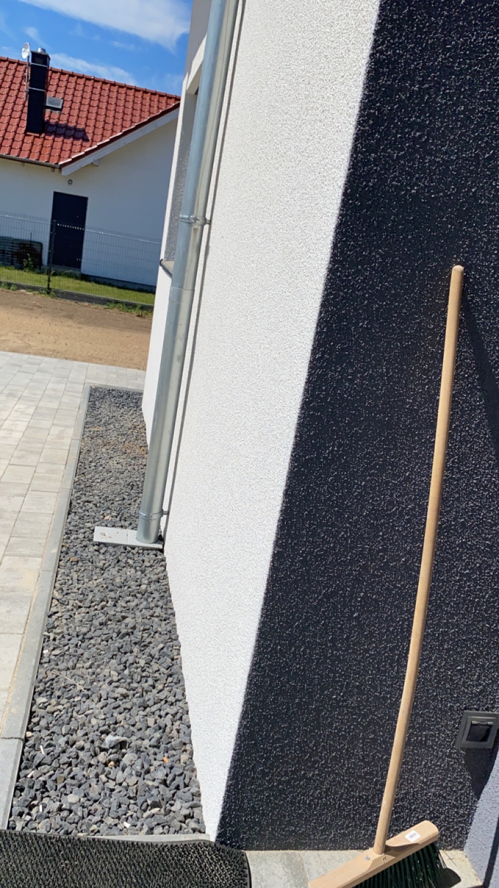 Fragment elewacji budynku z dwukolorową strukturą (biała i grafitowa) i rynną. Obok ściany leży miotła, na ziemi szare kamienie i wycieraczka.