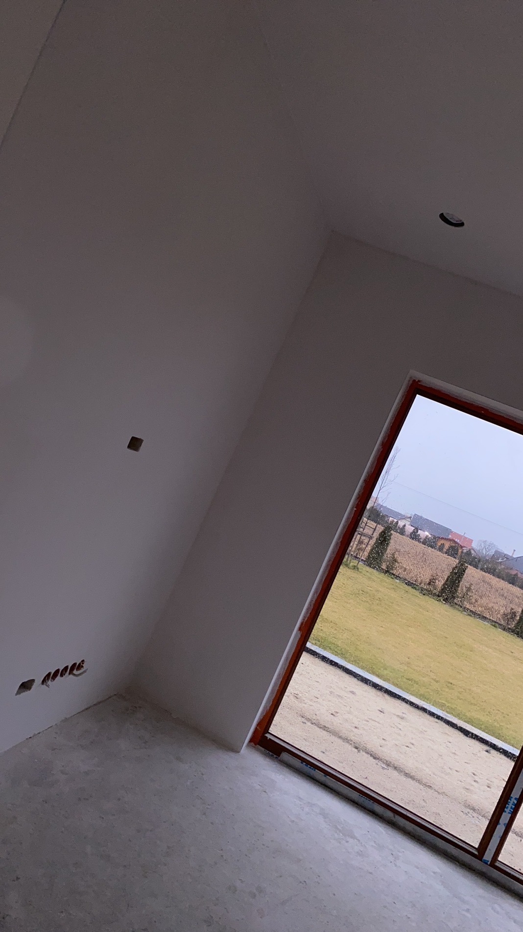 Surowe wnętrze z oknem na zewnątrz, widoczne przygotowania do wykończenia ścian i podłogi. Otwory na gniazdka elektryczne wskazują na etap prac instalacyjnych.