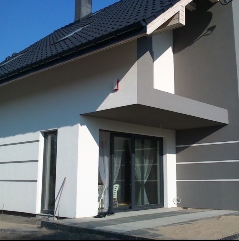 Elewacja domu po renowacji: białe ściany z szarymi akcentami, ciemny dach, nowoczesne okna i zadaszenie nad tarasem. Widoczny fragment ogrodu i schody wejściowe.