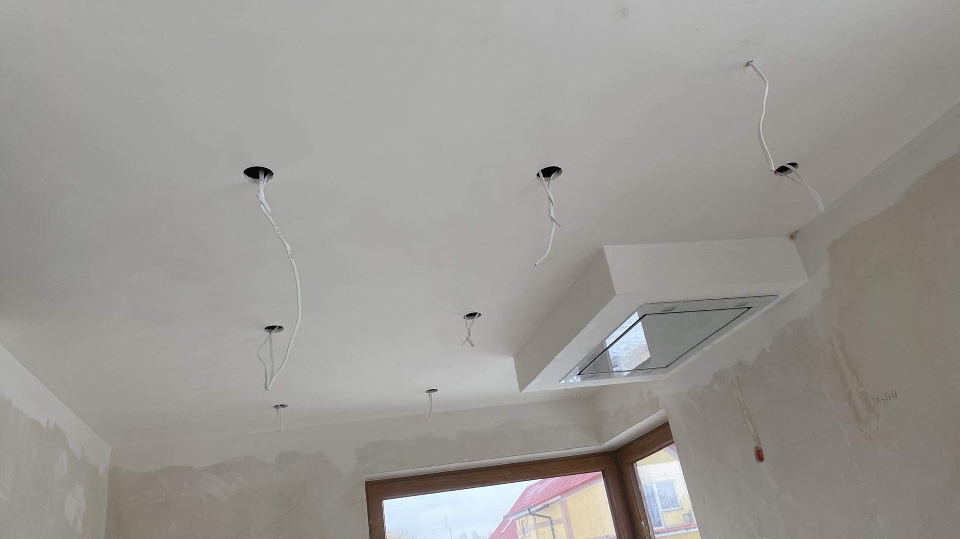 Białe wnętrze w trakcie wykończenia: surowy sufit z otworami na oświetlenie, wystające przewody, okno i fragment okapu kuchennego. Ściany przygotowane do malowania.