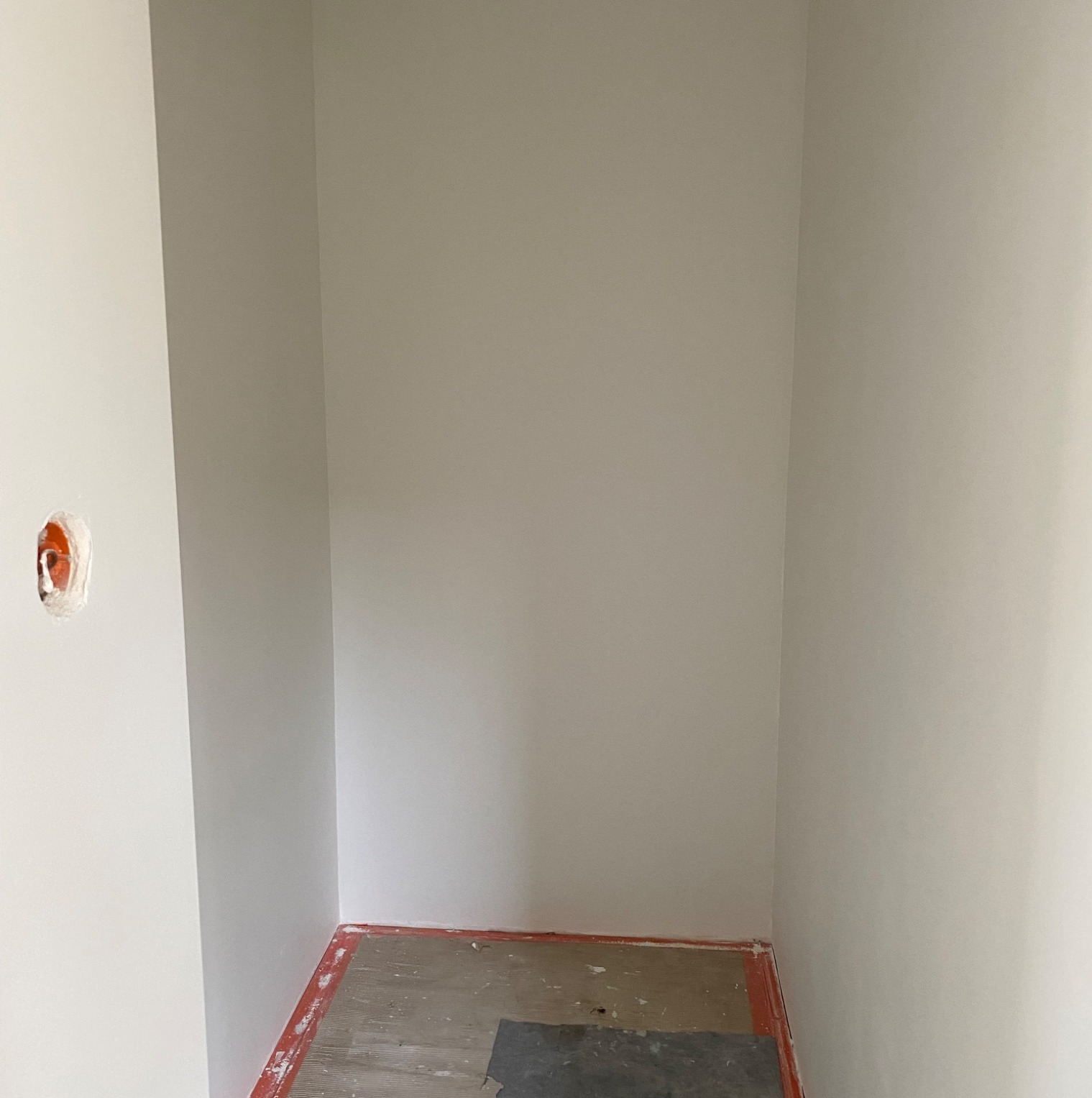 Wnętrze małego pomieszczenia po malowaniu ścian na jasny kolor, widoczne gniazdko elektryczne i fragment podłogi z pozostałościami po remoncie.