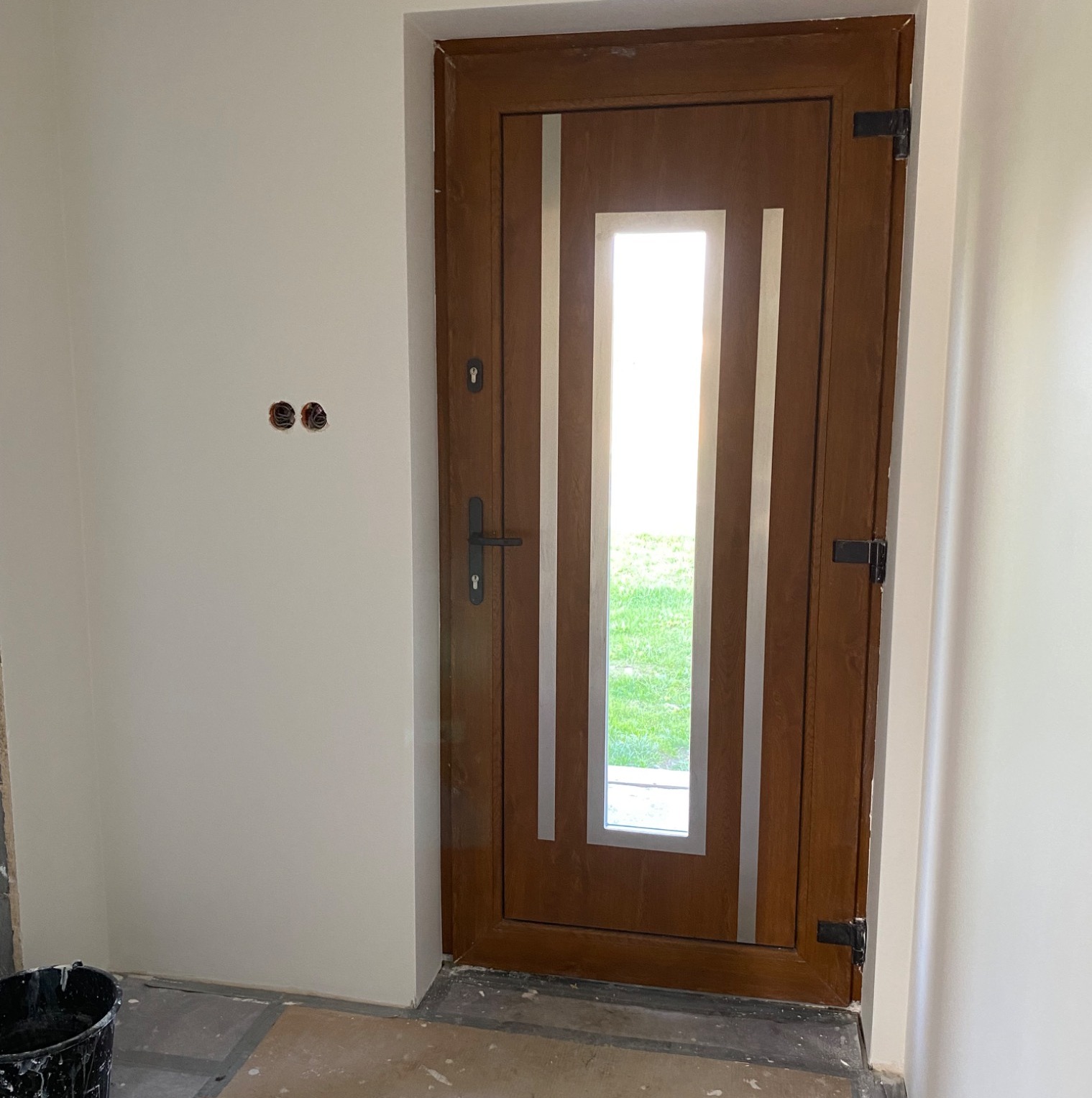 Brązowe drzwi wejściowe z szybą, osadzone w świeżo pomalowanej, jasnej ścianie. Widoczne ślady prac wykończeniowych na podłodze i puszki elektryczne bez osprzętu.