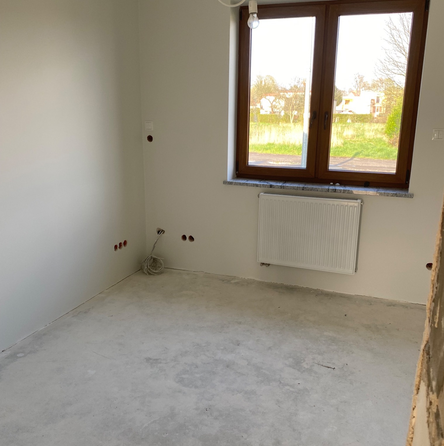 Surowe wnętrze pokoju z grzejnikiem pod oknem w drewnianej ramie. Widoczne wystające kable i niedokończona podłoga. Ściany pomalowane na biało.