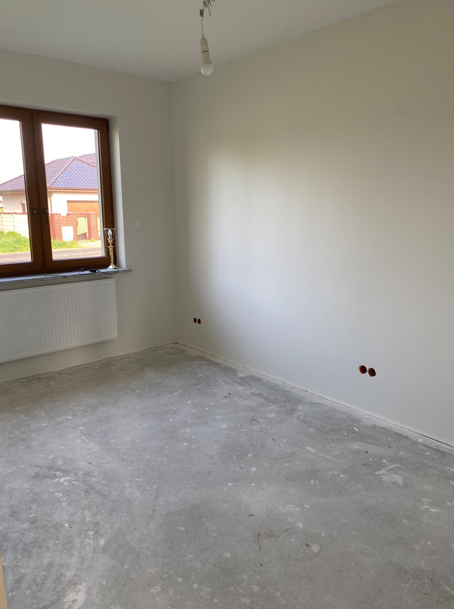 Surowe wnętrze pokoju z betonową podłogą, białymi ścianami i brązowym oknem. Widoczne gniazdka elektryczne i biały grzejnik pod oknem. W tle dom z czerwonym dachem.