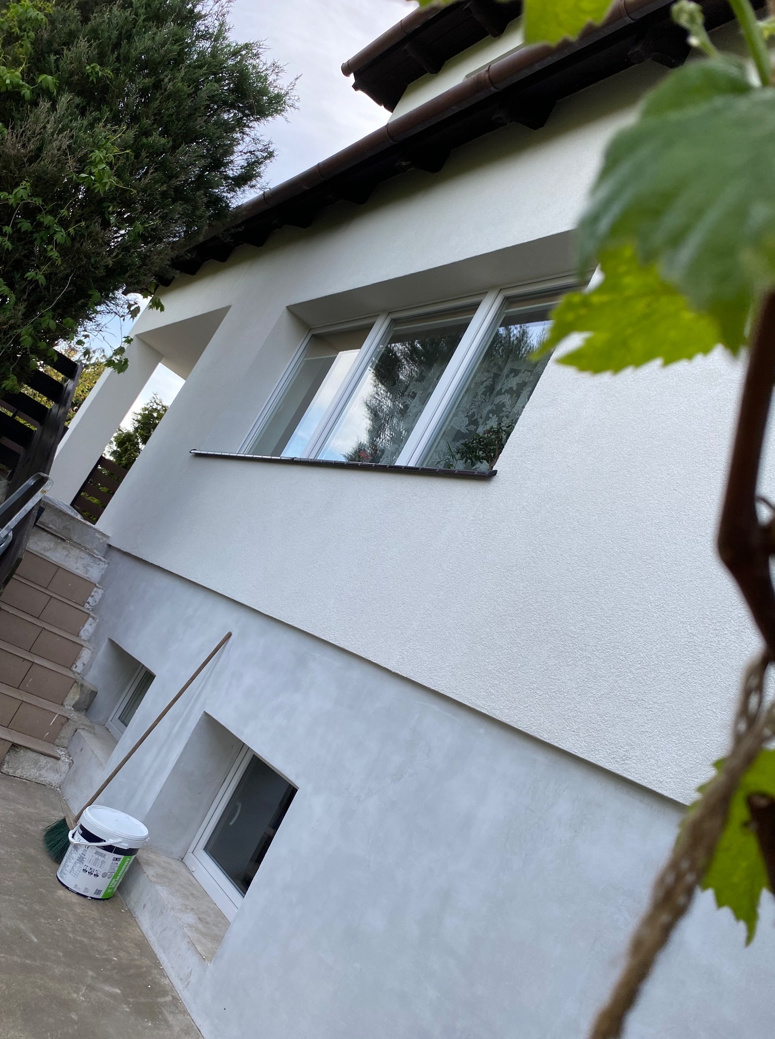 Ocieplony, biały budynek z widocznymi oknami, z boku widoczny pęd winorośli. Przy ścianie stoi wiadro z farbą i miotła. Świeżo pomalowana elewacja.