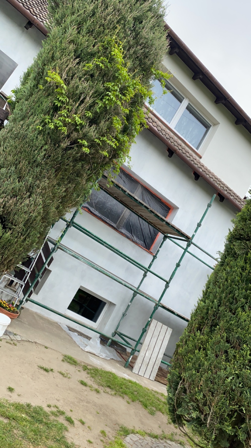 Dom w trakcie malowania elewacji. Okna zabezpieczone folią, rusztowanie przy ścianie. Widoczne deski i zieleń w tle. Świeżo pomalowana elewacja na biało.