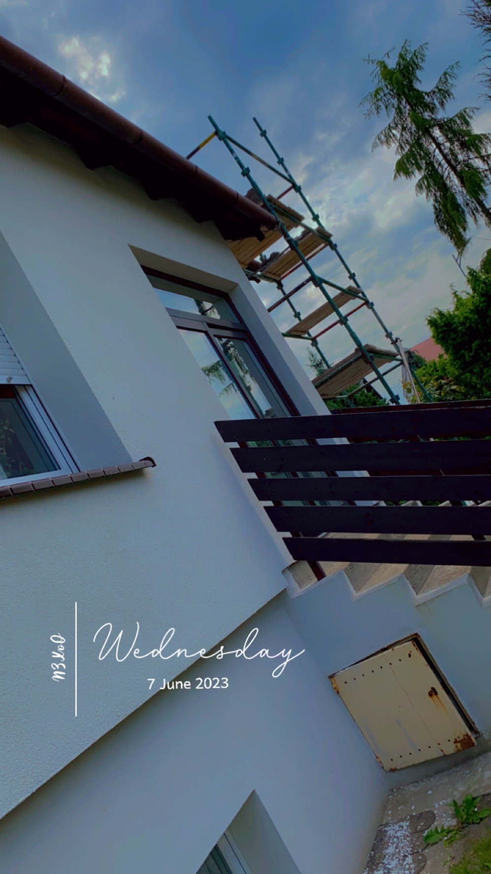 Biała elewacja domu z brązowym dachem i rusztowaniem. Widok pod kątem, fragment balkonu z drewnianą barierką. Częściowo widoczne okna i zieleń drzew.