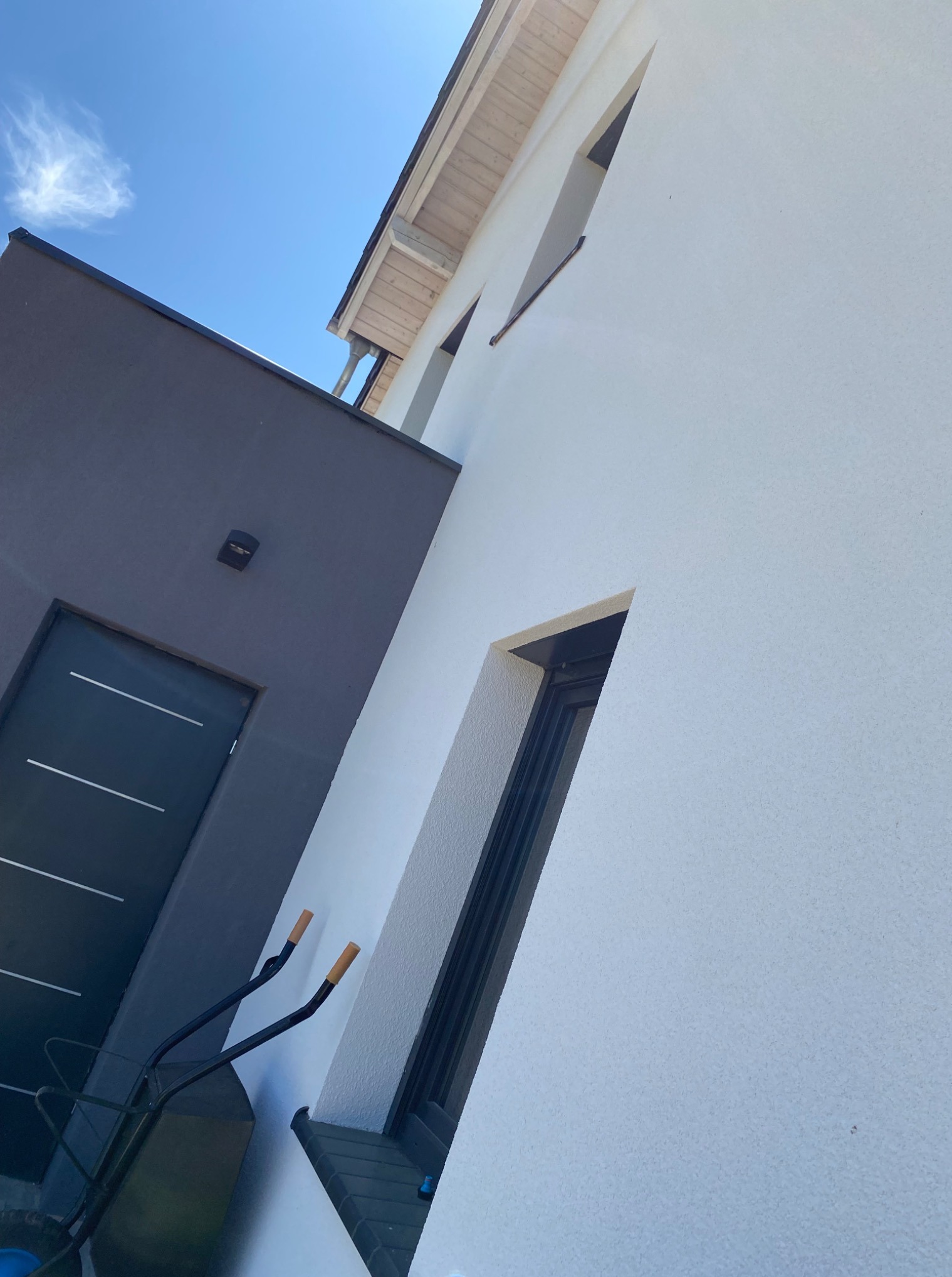 Biała elewacja domu z oknami i szarym wejściem, widok z dołu pod kątem. Taczka oparta o ścianę. Błękitne niebo w tle.
