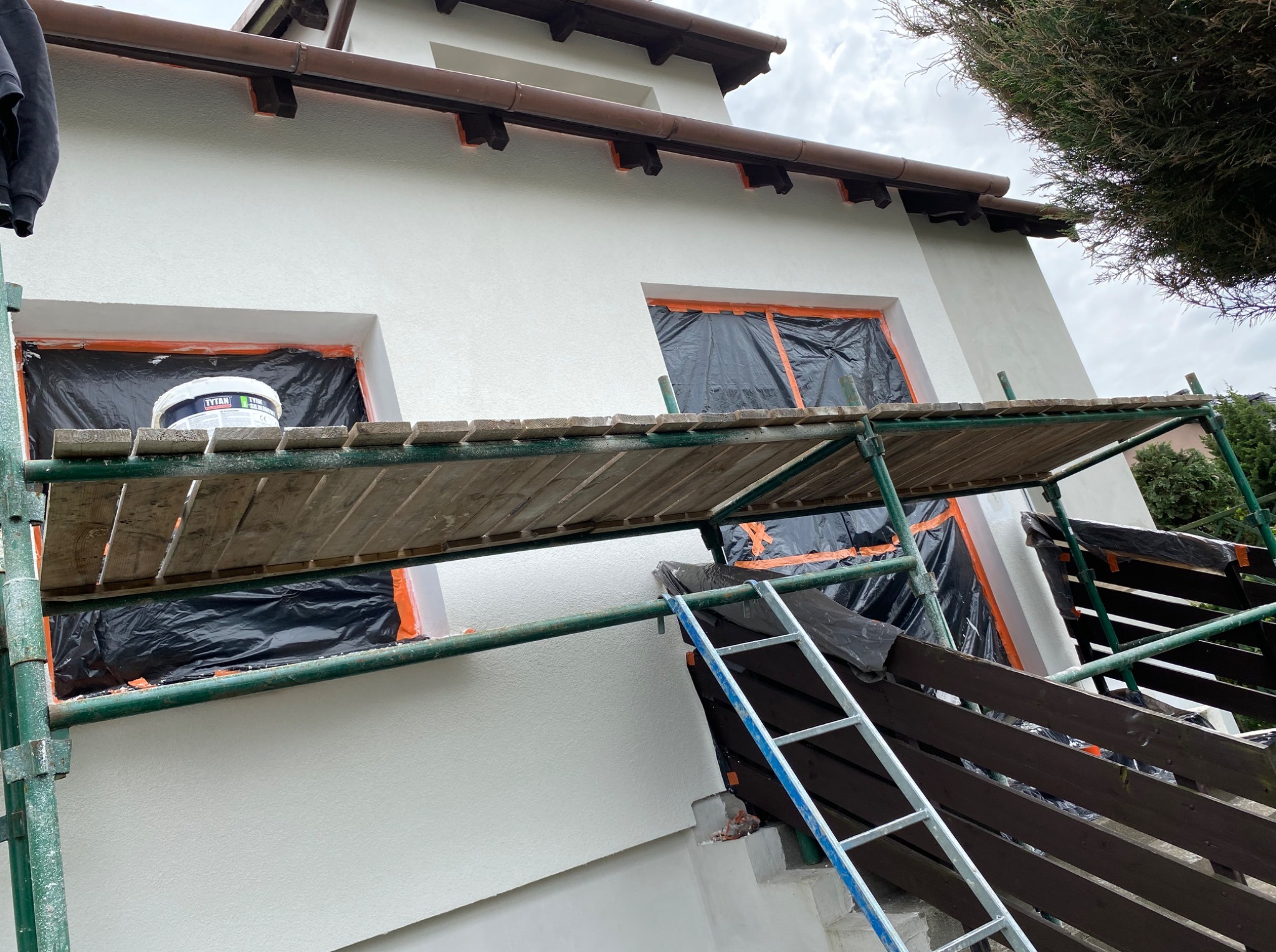 Biała fasada domu w trakcie malowania. Okna zabezpieczone folią, widoczne rusztowanie i drabina. Na rusztowaniu wiadro z farbą. Świeżo pomalowana elewacja.