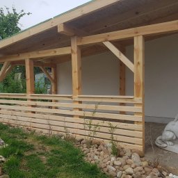 LukRem - Surowa drewniana konstrukcja zadaszenia tarasu z niską balustradą, widok z zewnątrz, z dekoracyjną figurą lwa i roślinnością w tle. Prace stolarskie.