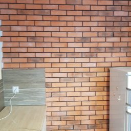 LukRem - Ściana z cegły klinkierowej w kuchni, widok na fragment blatu, gniazdko elektryczne i lodówkę. W tle kartony z elementami wykończeniowymi.