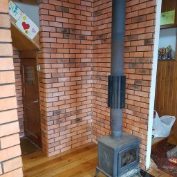 LukRem - Wykończenie wnętrza z ceglaną ścianą, drewnianą podłogą i piecem typu koza. Widoczny komin z metalową rurą. Dekoracja dziecięca na ścianie dodaje unikalnego charakteru.