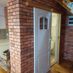 LukRem - Wykończenie wnętrza: ceglana ściana z przesuwnymi drzwiami do łazienki, drewniana podłoga. Widoczne fragmenty drabiny i kartonów.