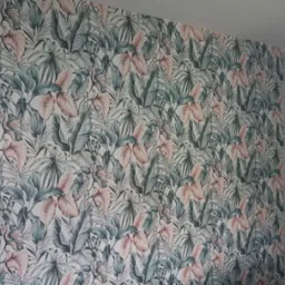 Ściana pokryta tapetą w tropikalne liście w odcieniach zieleni i różu, widoczny fragment szarej ściany i ościeżnica drzwi. Ukończone tapetowanie.