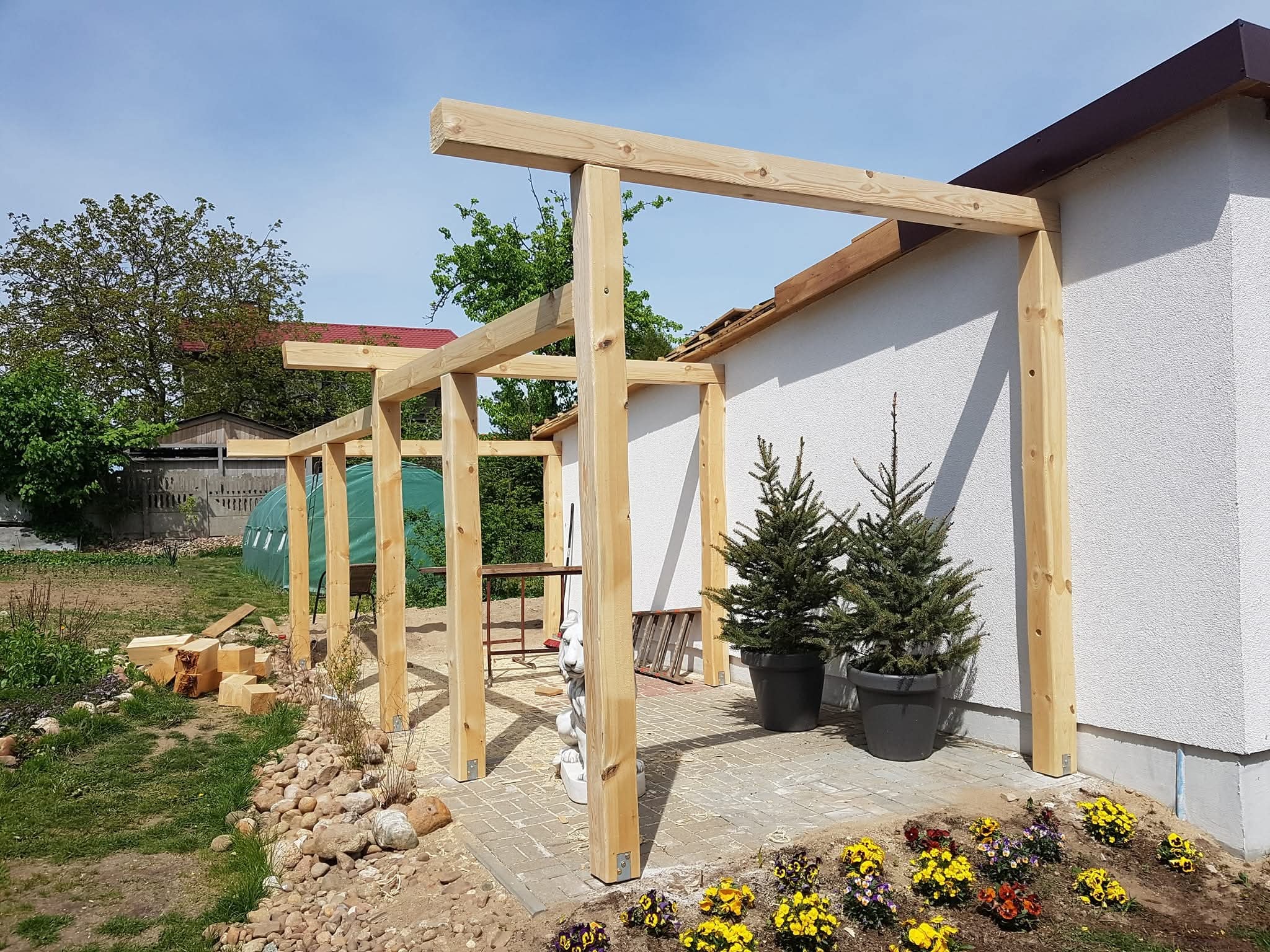 Drewniana konstrukcja z belek, prawdopodobnie pergola, przymocowana do białego budynku. Widoczne kwiaty, tuje w donicach i elementy krajobrazu.
