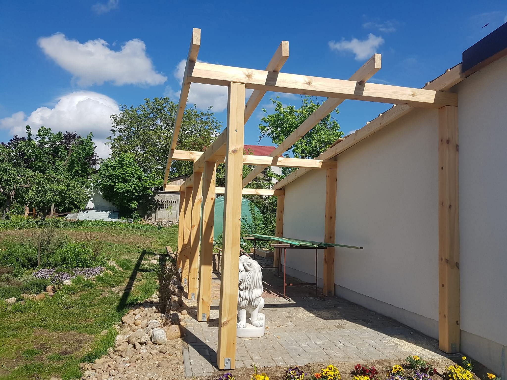 Surowa drewniana konstrukcja pergoli przy białej ścianie domu, z widocznym ogrodem i rzeźbą lwa. Słoneczny dzień, błękitne niebo z chmurami.