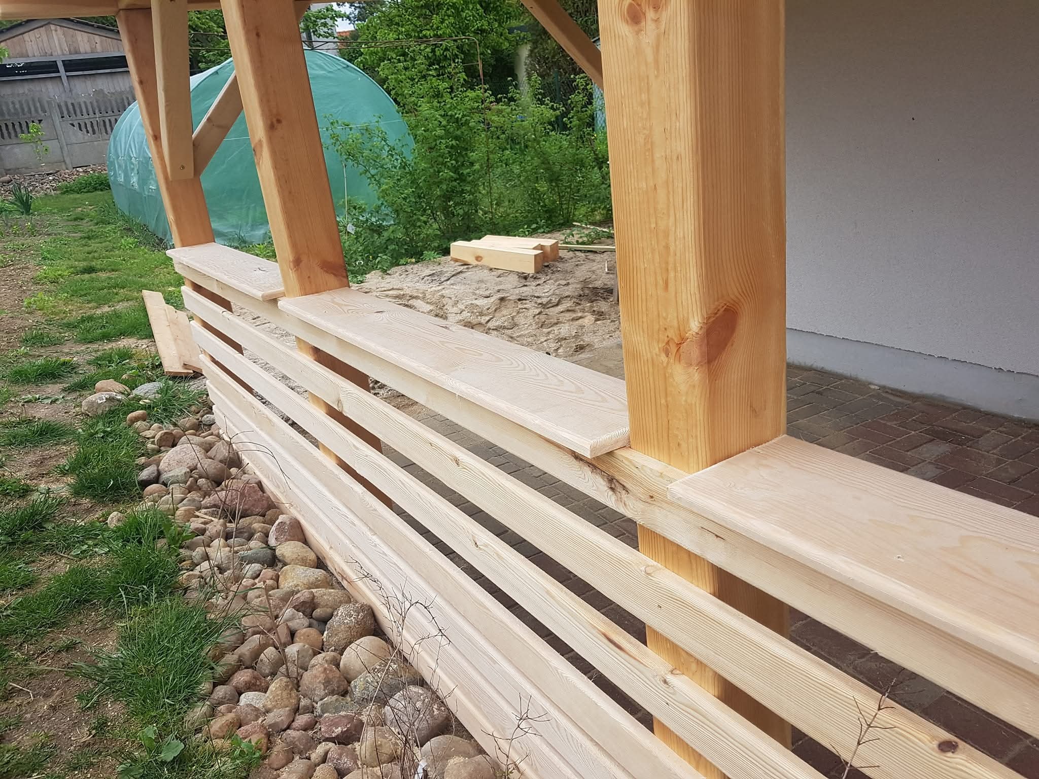 Drewniana konstrukcja z bali i desek, prawdopodobnie altana lub pergola w trakcie budowy. Widoczne elementy konstrukcyjne i fragment ogrodu w tle.
