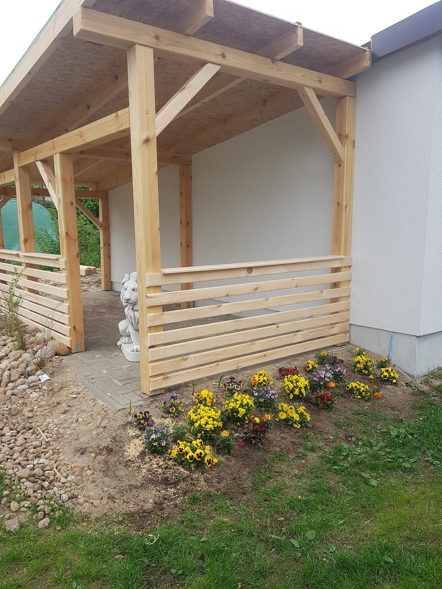 Drewniana altana z płotkiem, ozdobiona kwiatami i figurą lwa. Nowoczesny design z jasnego drewna, zadaszona, przylega do białego budynku. Urokliwa aranżacja.