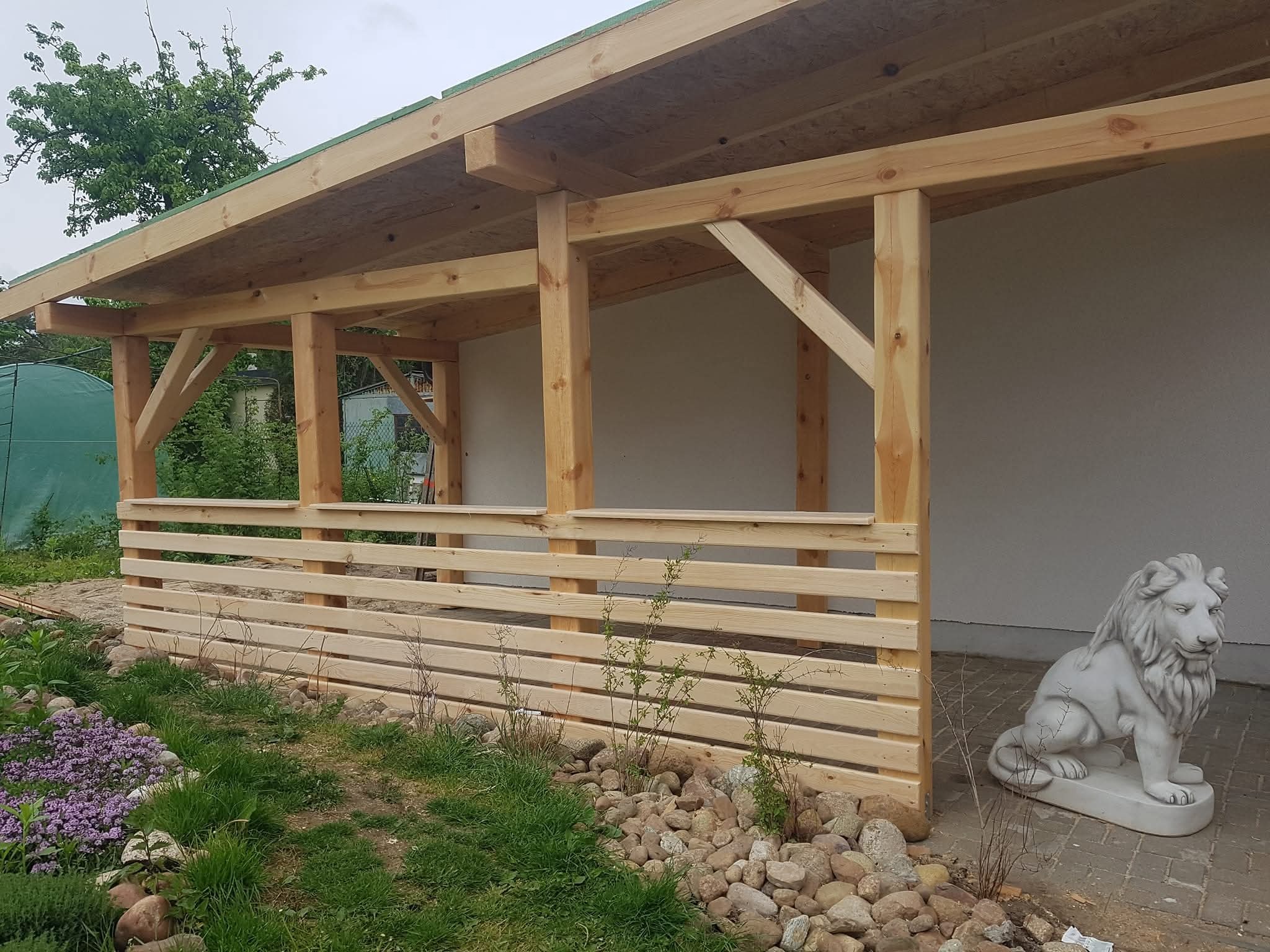 Surowa drewniana konstrukcja zadaszenia tarasu z niską balustradą, widok z zewnątrz, z dekoracyjną figurą lwa i roślinnością w tle. Prace stolarskie.