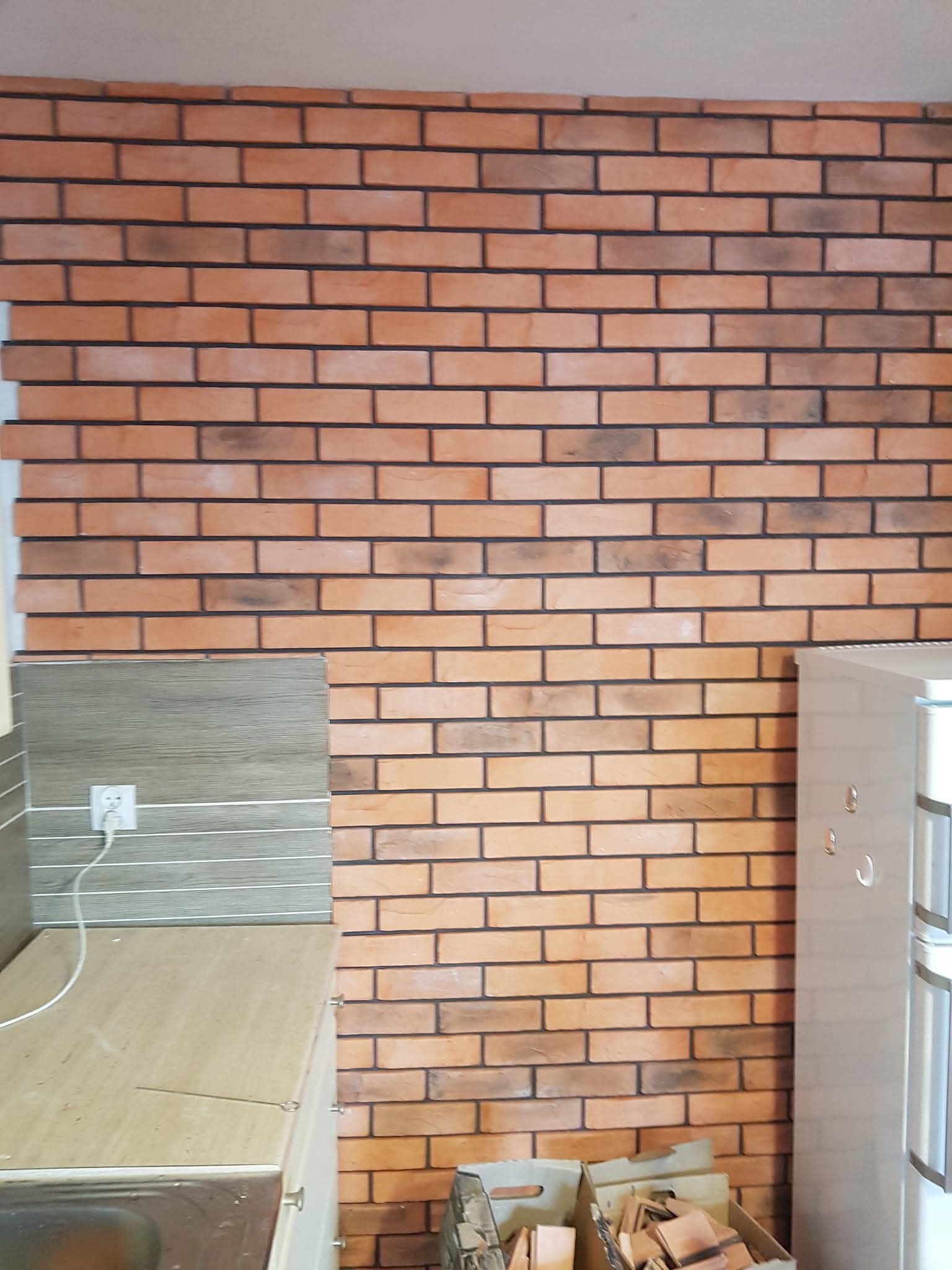 Ściana z cegły klinkierowej w kuchni, widok na fragment blatu, gniazdko elektryczne i lodówkę. W tle kartony z elementami wykończeniowymi.