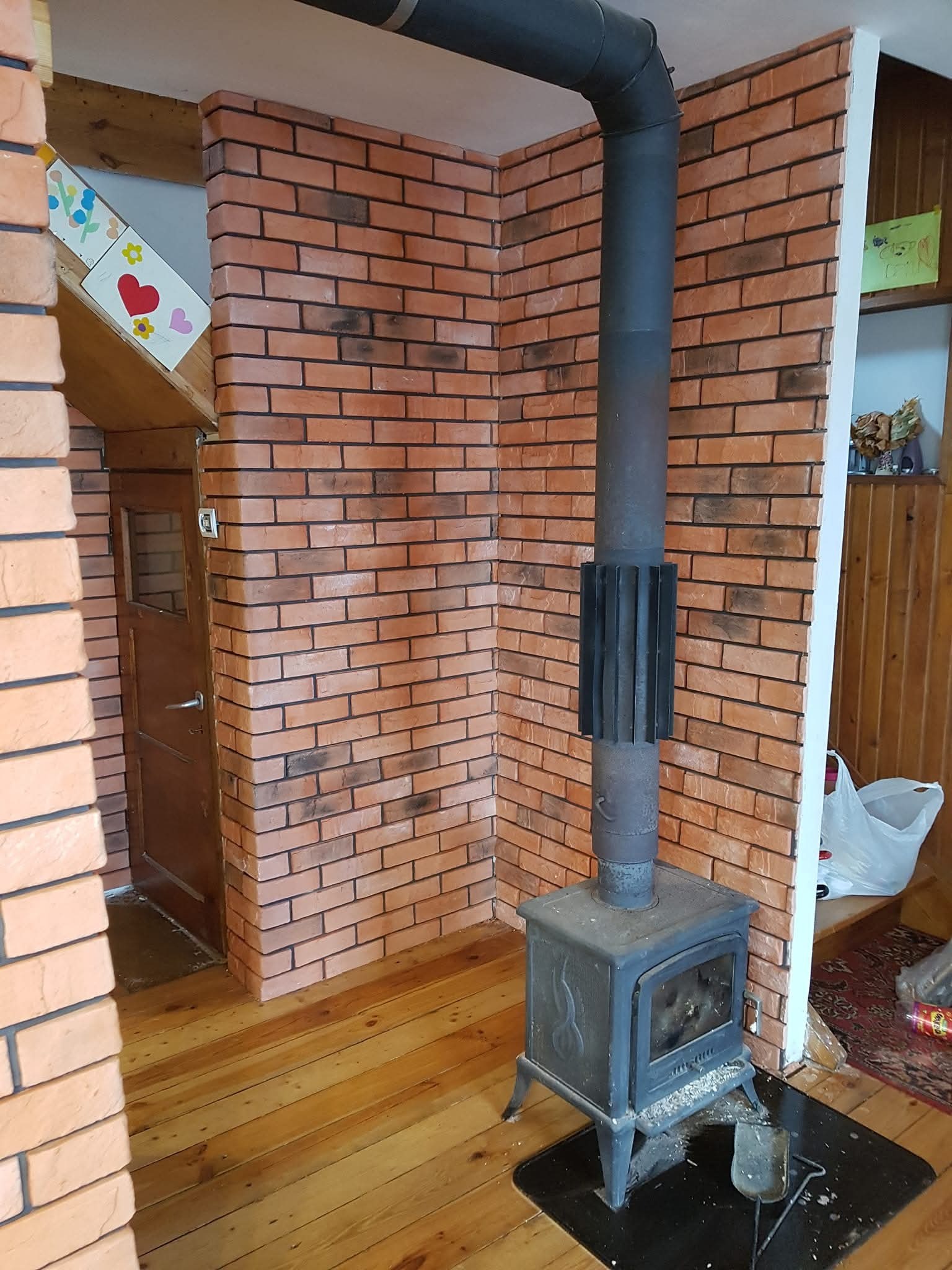 Wykończenie wnętrza z ceglaną ścianą, drewnianą podłogą i piecem typu koza. Widoczny komin z metalową rurą. Dekoracja dziecięca na ścianie dodaje unikalnego charakteru.