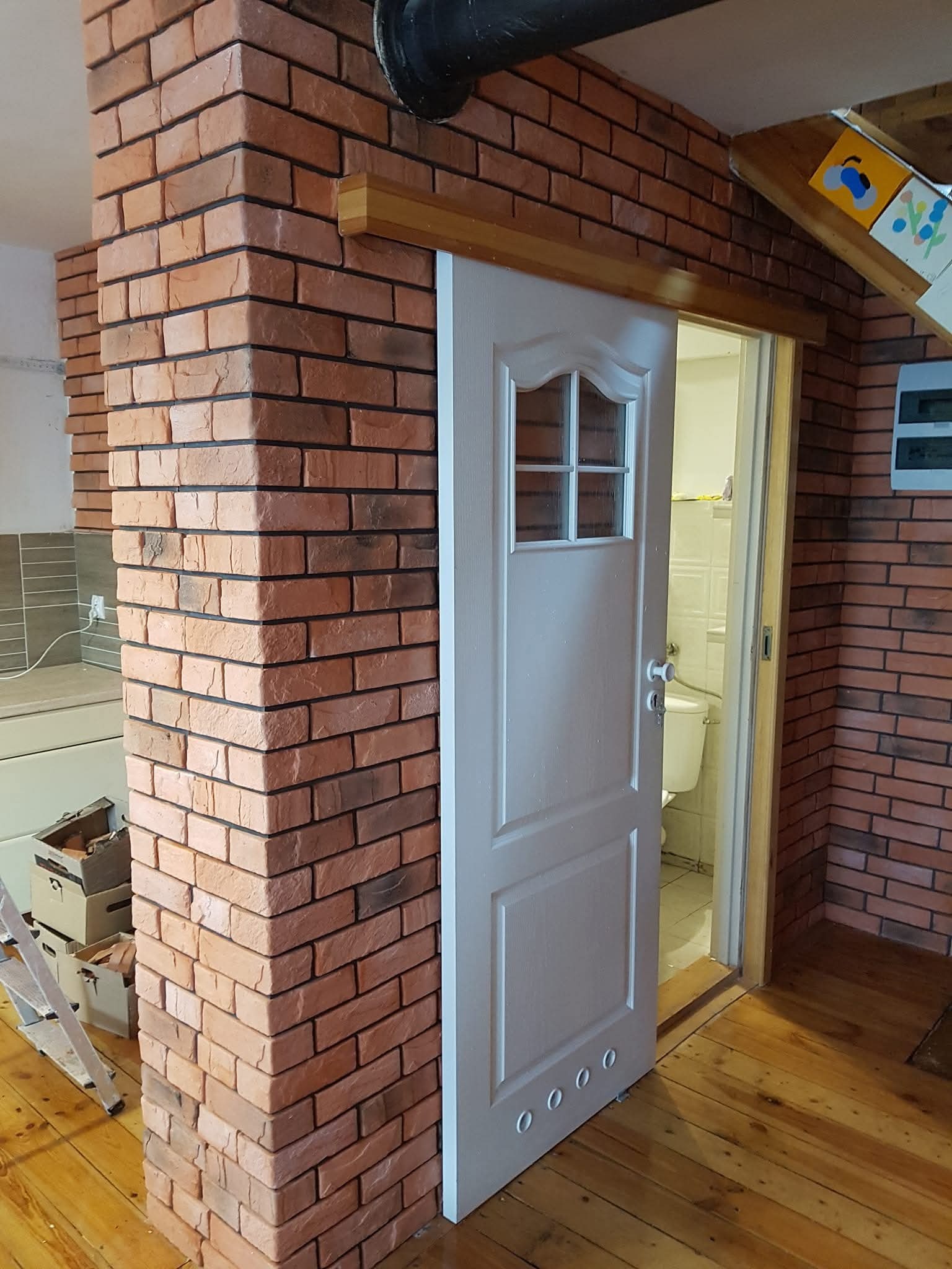 Wykończenie wnętrza: ceglana ściana z przesuwnymi drzwiami do łazienki, drewniana podłoga. Widoczne fragmenty drabiny i kartonów.