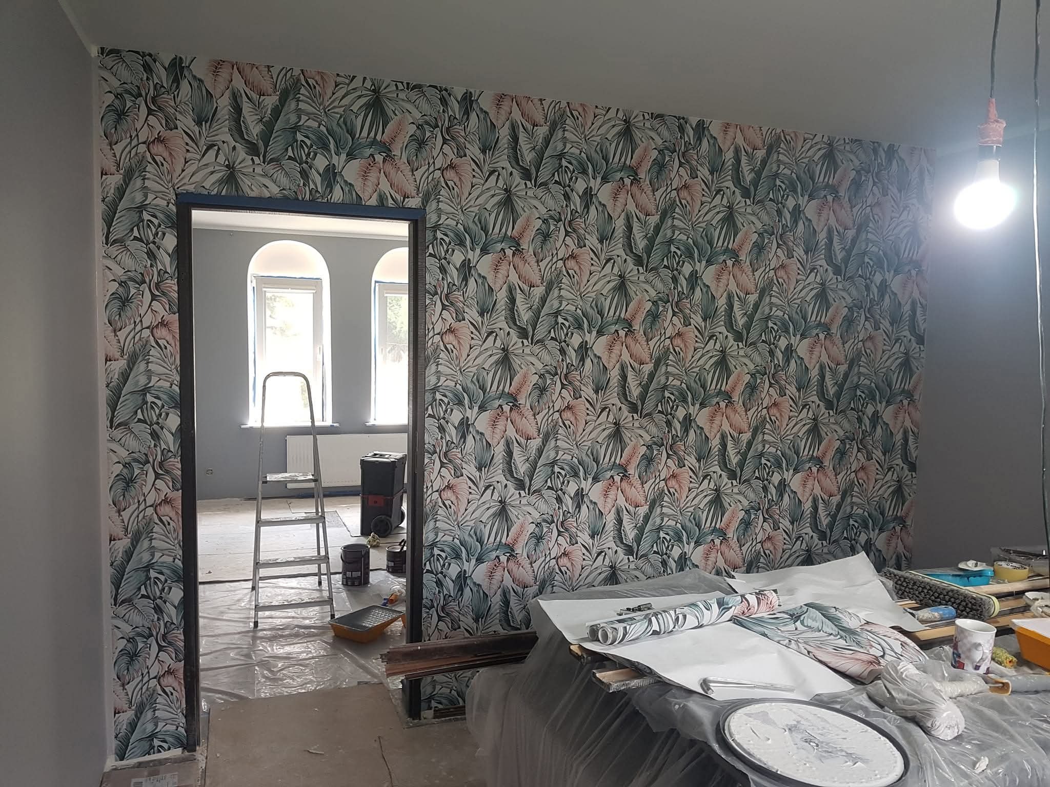 Ściana z tapetą w tropikalne liście, widok przez drzwi na pokój ze schodami malarskimi. W pomieszczeniu narzędzia malarskie i rolki tapety.