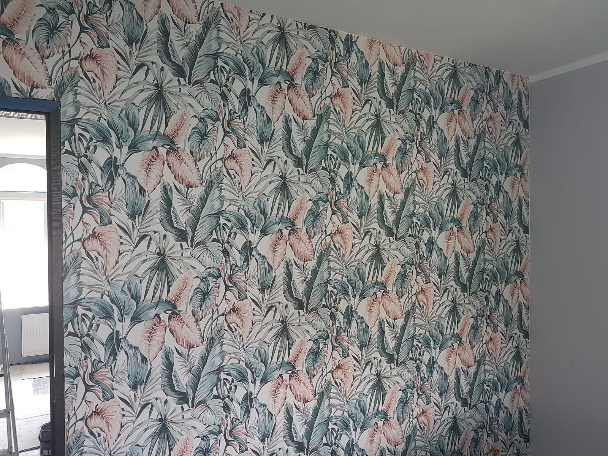 Ściana pokryta tapetą w tropikalne liście w odcieniach zieleni i różu, widoczny fragment szarej ściany i ościeżnica drzwi. Ukończone tapetowanie.