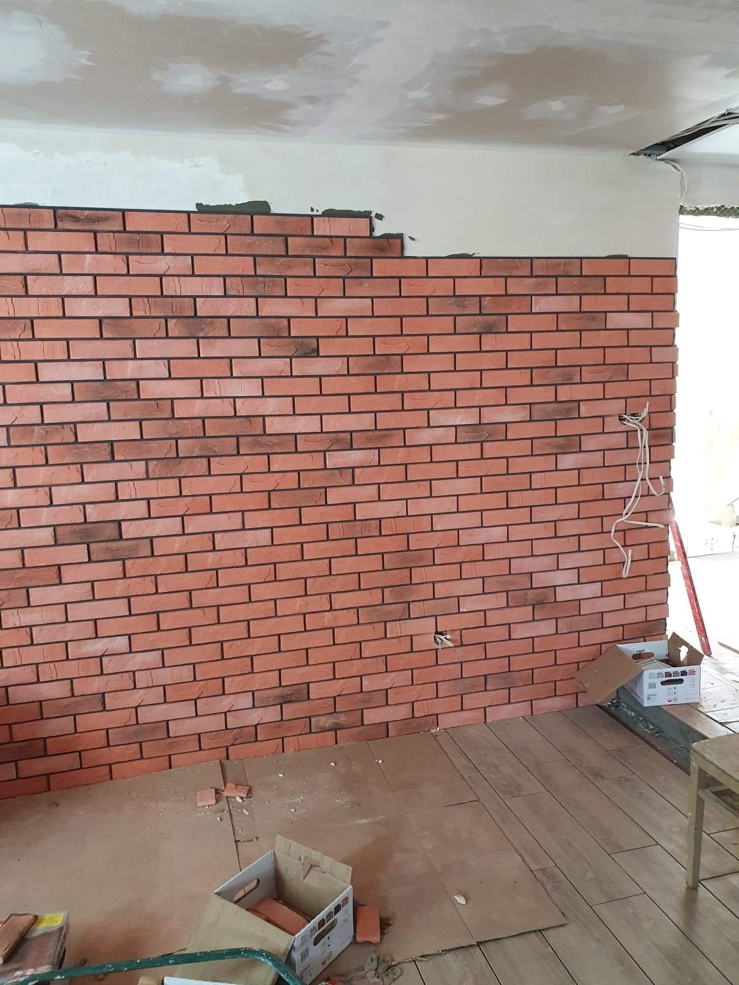 Układanie cegieł dekoracyjnych na ścianie, widoczne prace wykończeniowe, częściowo ułożona ściana, podłoga z drewnopodobnych paneli, kartony z cegłami.