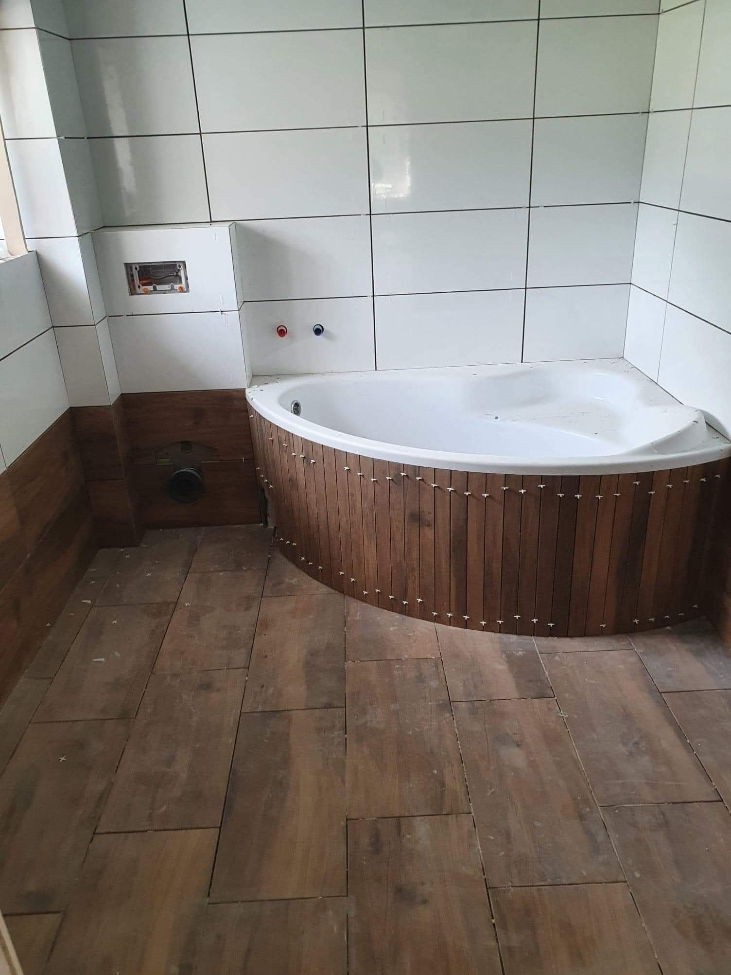 Narożna wanna z obudową z drewnopodobnych paneli w trakcie montażu. Widoczne płytki na podłodze i ścianach, białe i drewnopodobne. Częściowo zamontowana armatura.