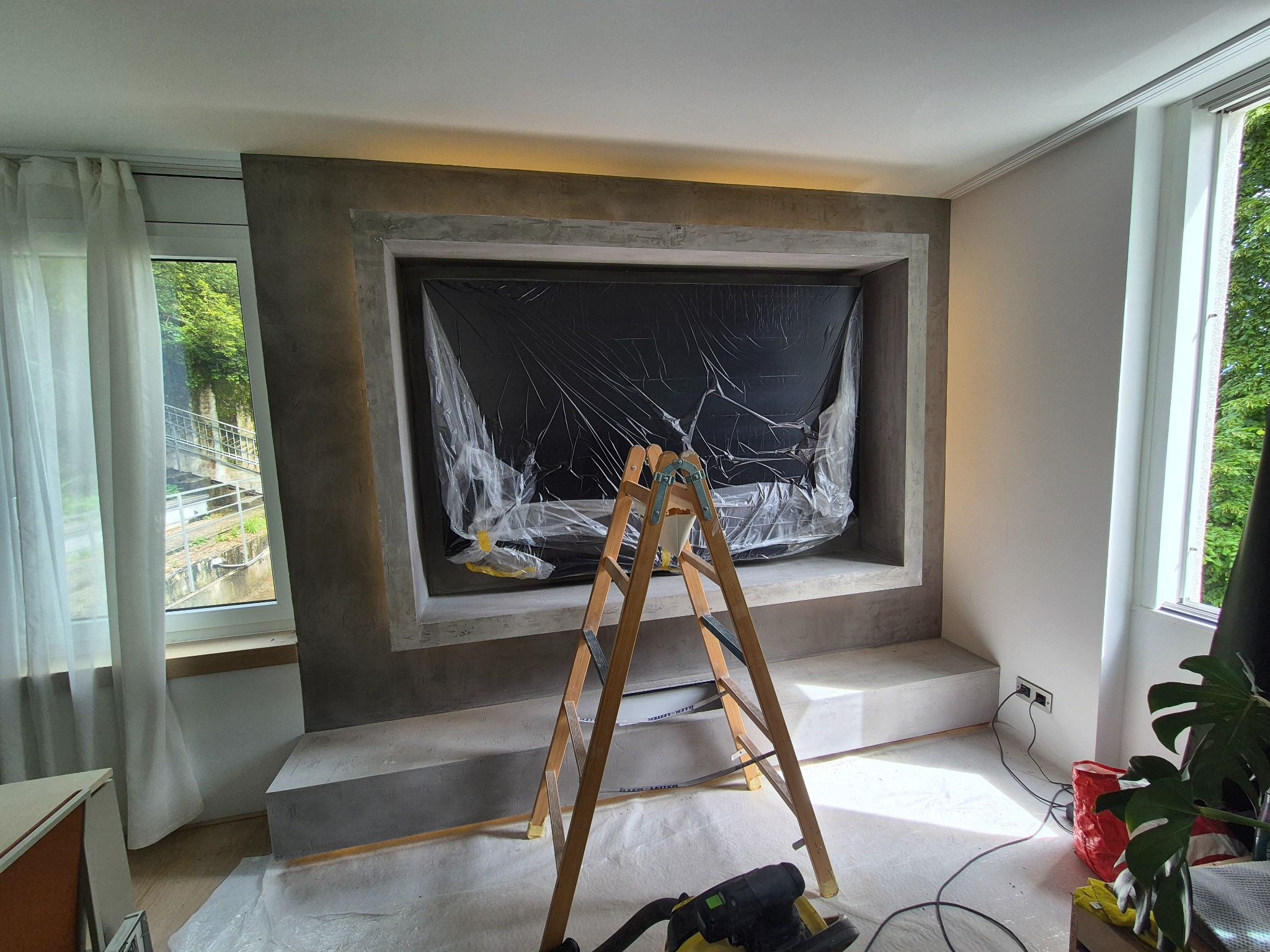 Pokój w trakcie prac: drabina stoi przed obudową TV zabezpieczoną folią, widoczny odkurzacz i okno z widokiem na zieleń. Przygotowania do dalszych prac.