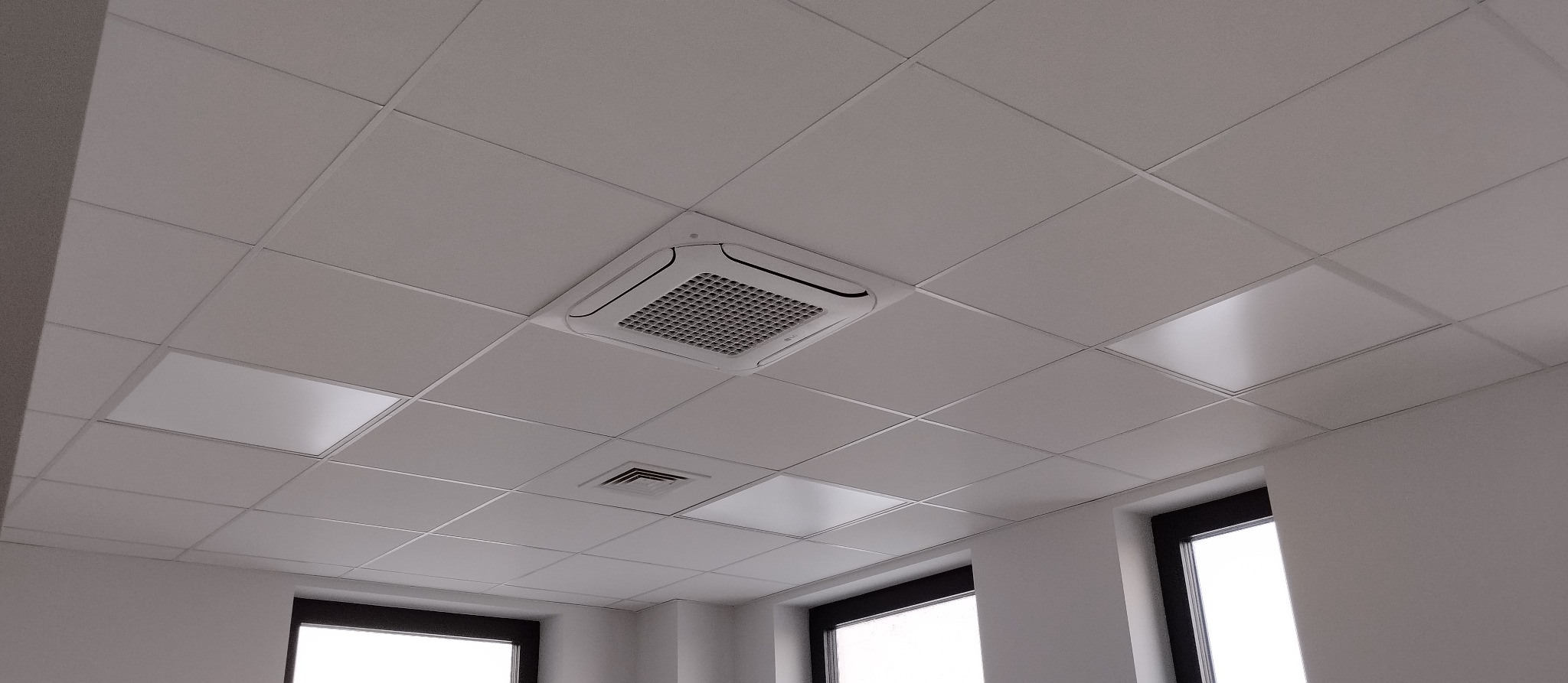 Biała kaseta klimatyzacyjna sufitowa w biurze, wkomponowana w kasetonowy sufit podwieszany. Widoczne okna z czarną ramą i kratka wentylacyjna.