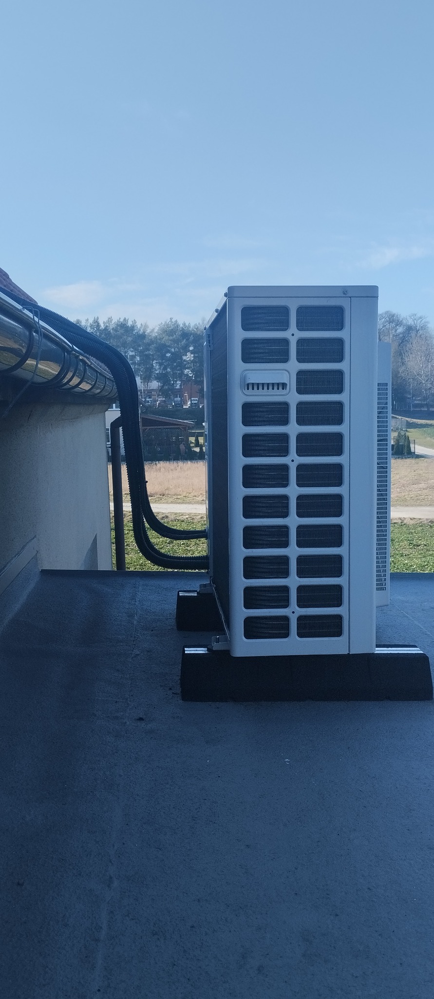 Agregat klimatyzacji kanałowej na dachu wiatrołapu, z rurami w peszlu UV. Widok z boku, na tle drzew i błękitnego nieba. Zabezpieczenia na rurach.