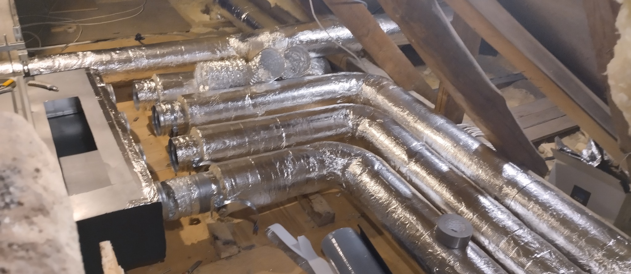Kanały wentylacyjne w izolacji aluminiowej, zamontowane pod skosem dachu, widoczne narzędzia i elementy montażowe. Etap instalacji przed ociepleniem puszki rozprężnej.