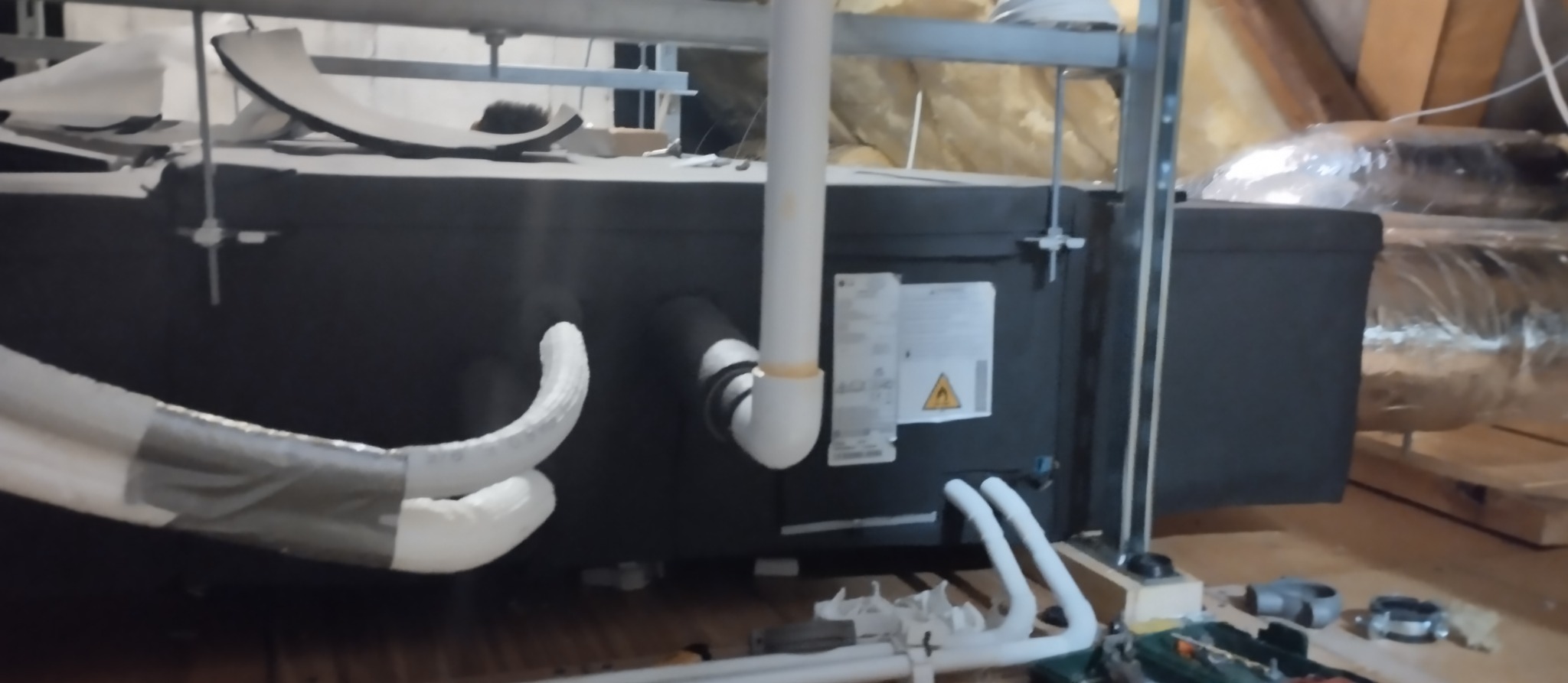 Czarna centrala wentylacyjna z rurami, zamontowana na poddaszu z widocznymi elementami konstrukcyjnymi dachu i narzędziami. Widoczny fragment instalacji.