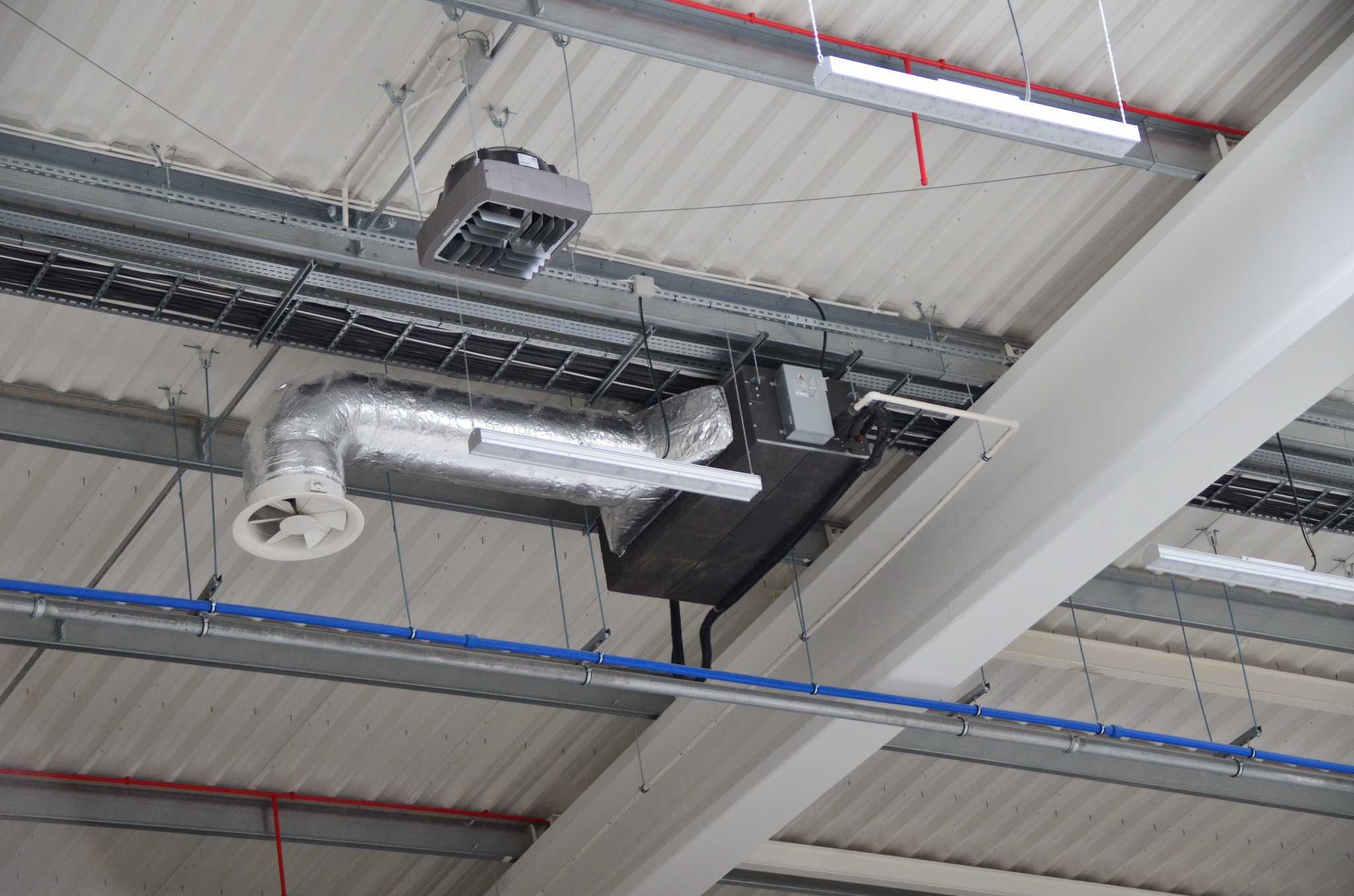 Instalacja wentylacyjna pod sufitem hali: srebrne rury, biały anemostat, czarna skrzynka z przewodami, oświetlenie LED i niebieskie rury. Widok z dołu na system.