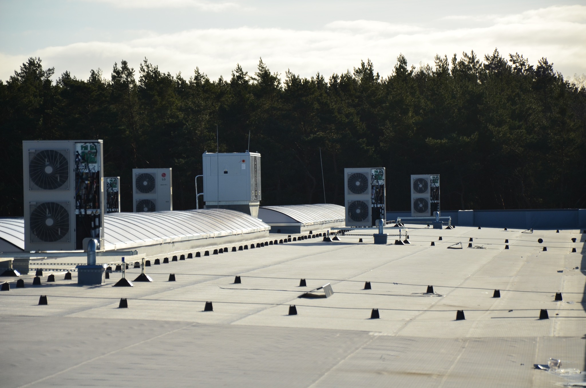 Instalacja klimatyzacji na dachu budynku z widokiem na las w tle. Widoczne jednostki zewnętrzne LG i elementy mocujące. Jasne niebo z chmurami.