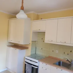 Jasna kuchnia z kremowymi szafkami, kafelkami z motywem koguta i lampą w stylu vintage. Ujęcie prezentuje wnętrze po remoncie, ukazując detale wykończenia.