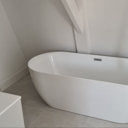 Kamil Mikołajski usługi ogólnobudowlane - Nowoczesna, wolnostojąca wanna w minimalistycznej łazience z szarą podłogą i białymi ścianami. Uwagę zwraca ukośny element architektoniczny na suficie.